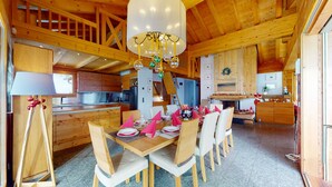 A spacious wooden dining area with a high ceiling, a chandelier, and a kitchen in the background.