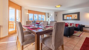 A dining area with a wooden table set for four, a sofa, and a view of mountains through large windows. - Myosotis 10 - QUIET apartment 8 pers (Veysonnaz)