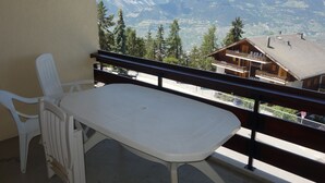 A balcony with a white table and chairs, offering a view of a mountain landscape and a wooden building.