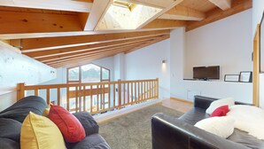 Un vaste espace de vie doté d’un plafond haut et de poutres en bois, un balcon avec une balustrade en bois, un canapé en cuir noir, un plaid en fausse fourrure blanche et un téléviseur fixé au mur.