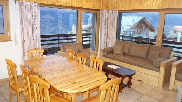 A wooden-furnished living room with a dining table, sofa, and large windows offering mountain views. - Mont-Rouge E 010 - VIEW apartment 8 pers (Veysonnaz)