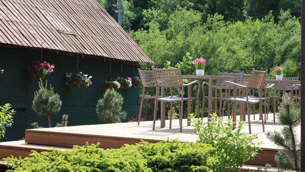 An outdoor patio with wooden chairs and tables, surrounded by potted plants and a green building with a sloped roof. - Beautiful modern villa with large suites in the middle of the mountains (Oščadnica )