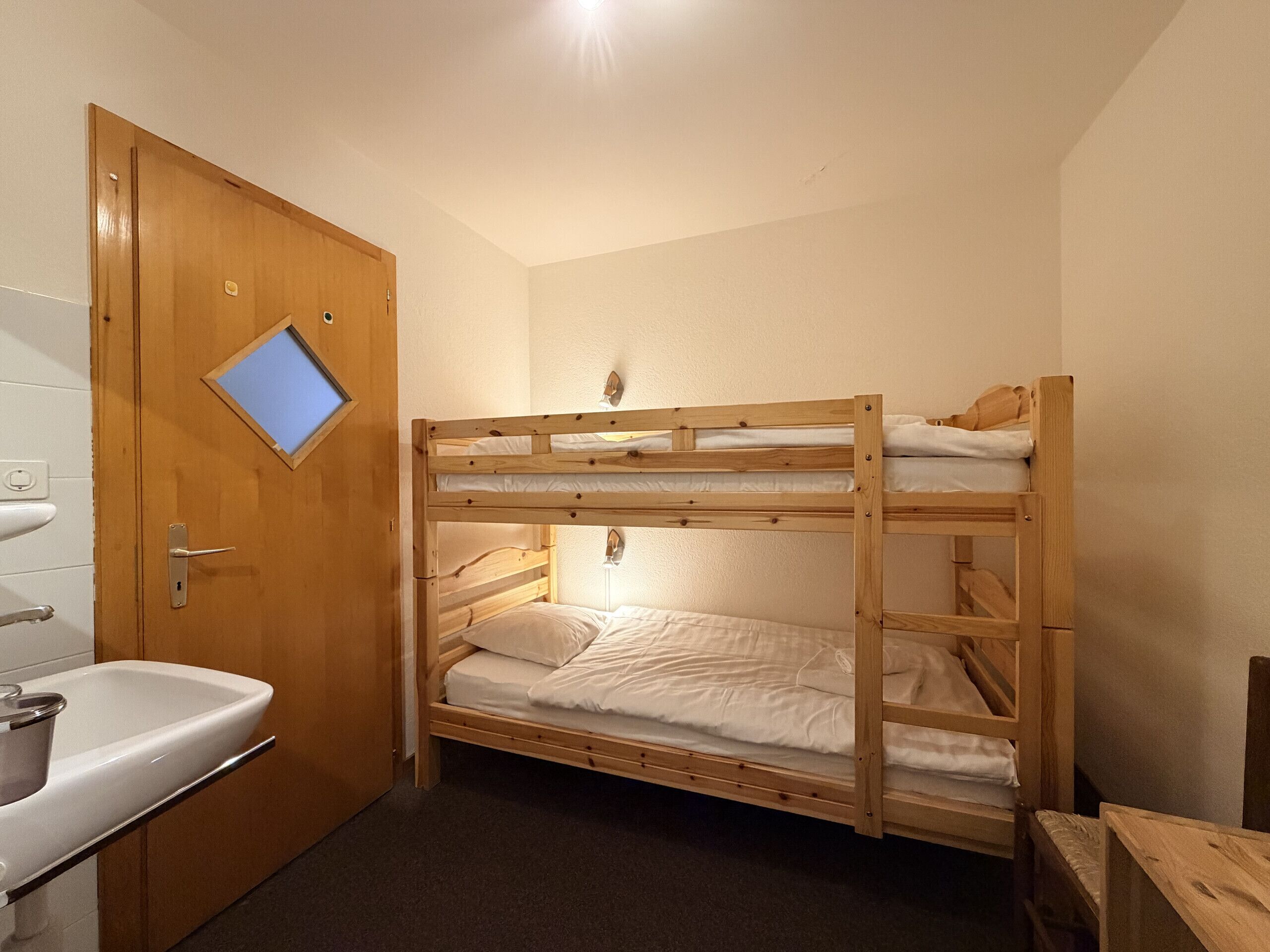 Een slaapkamer met een stapelbed, een houten deur, een klein raam en een wastafel.