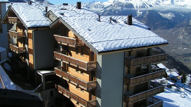 A multi-story wooden building with balconies, set against a backdrop of snow-covered mountains.