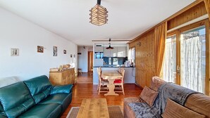 A living room with a green sofa, a wooden coffee table, and a dining area with a wooden table and chairs. There is a kitchen in the background with white cabinets and a black ceiling fan. - Iris I 023 - CONFORTABLE apartment 8 pers (Veysonnaz)