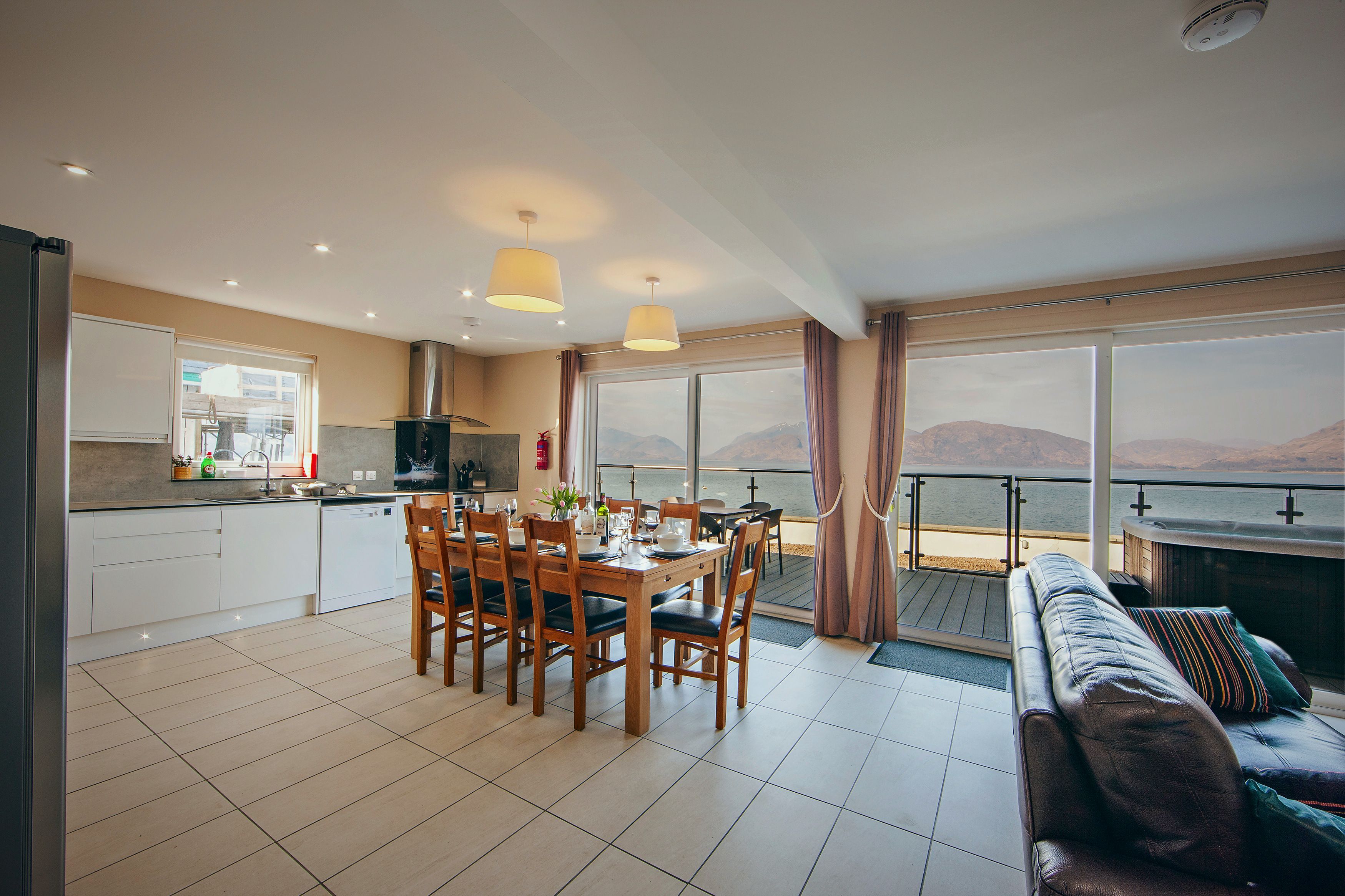 A modern kitchen with a dining area, large windows, and a balcony overlooking a body of water.
