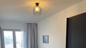Ein kleiner, ordentlicher Raum mit einem Bett, einem Vorhangfenster, einer Wandleuchte und einer Tür.