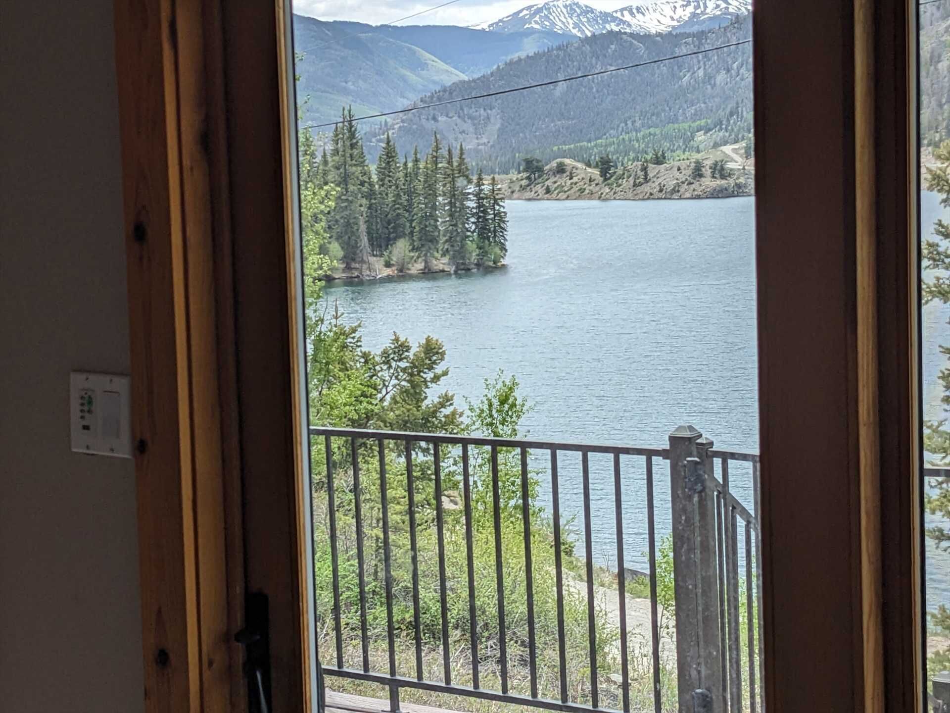 A view from a balcony with a metal railing, overlooking a lake surrounded by trees and mountains.