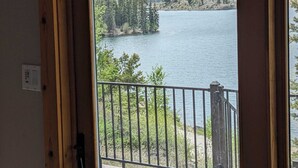 Blick von einem Balkon mit Metallgeländer auf einen See, umgeben von Bäumen und Bergen.