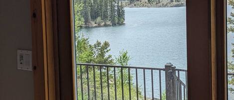 A view from a balcony with a metal railing, overlooking a lake surrounded by trees and mountains.
