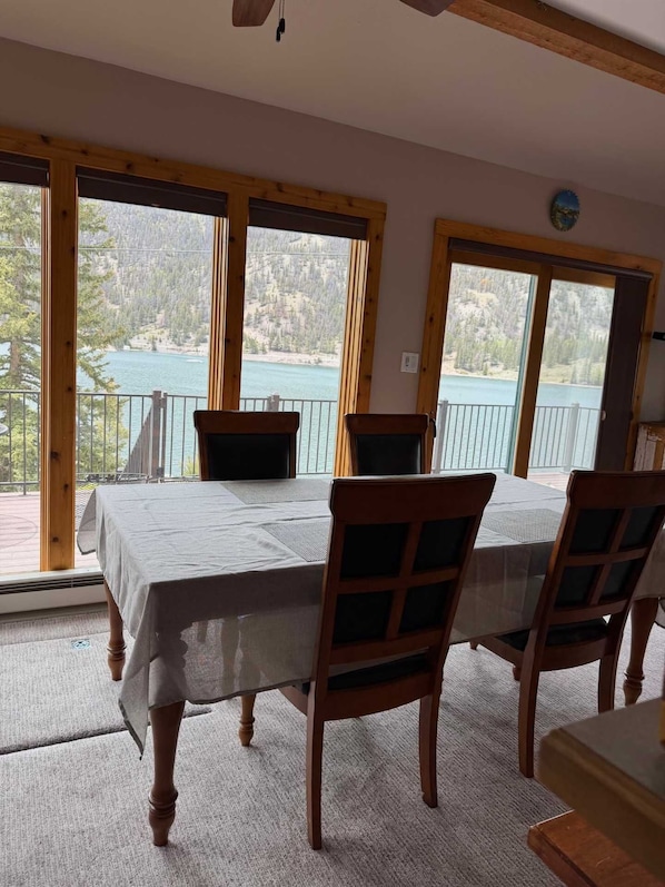 Et spiseområde med et bord dækket til fire personer, store vinduer med udsigt over sø og skov, og træbjælker i loftet.