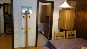Una habitación con suelo de madera, una mesa con mantel, perchero de madera, armario de madera y una cocina visible a través de una puerta abierta.