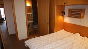 Un dormitorio con cama, cabecero de madera, ventana con persianas y un baño visible a través de una puerta abierta.