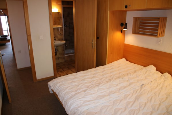 A bedroom with a bed, a wooden headboard, a window with blinds, and a bathroom visible through an open door.