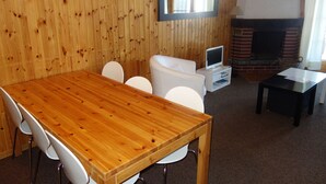 Een kamer met houten lambrisering, een houten eettafel, witte stoelen, een witte bank, een televisie en een open haard.