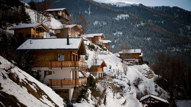 Een groep houten chalets op een besneeuwde helling, met een berg op de achtergrond.