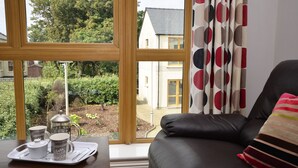 Een gezellige woonkamer met een leren bank, een raam met uitzicht op een tuin en een huis, en een dienblad met koffiekopjes en een theepot op een houten tafel.