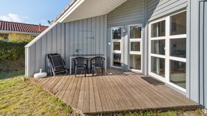 Una terraza de madera con una mesa y sillas, un portón corredero de vidrio y un pequeño espacio exterior con una silla y una escalera.