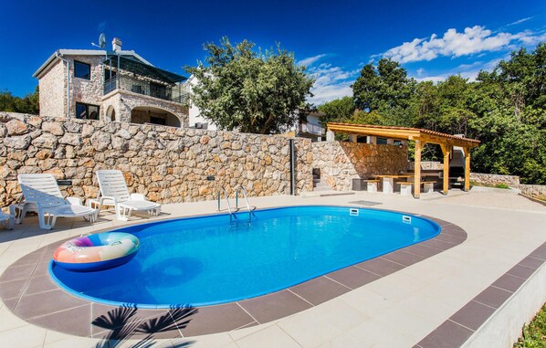 Relax by the pool surrounded by lush greenery and lounge chairs, with a barbecue area nearby.