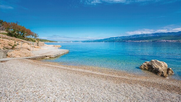 Een kiezelstrand met helderblauw water, een betonnen pad en rotsachtig terrein met schaarse begroeiing.