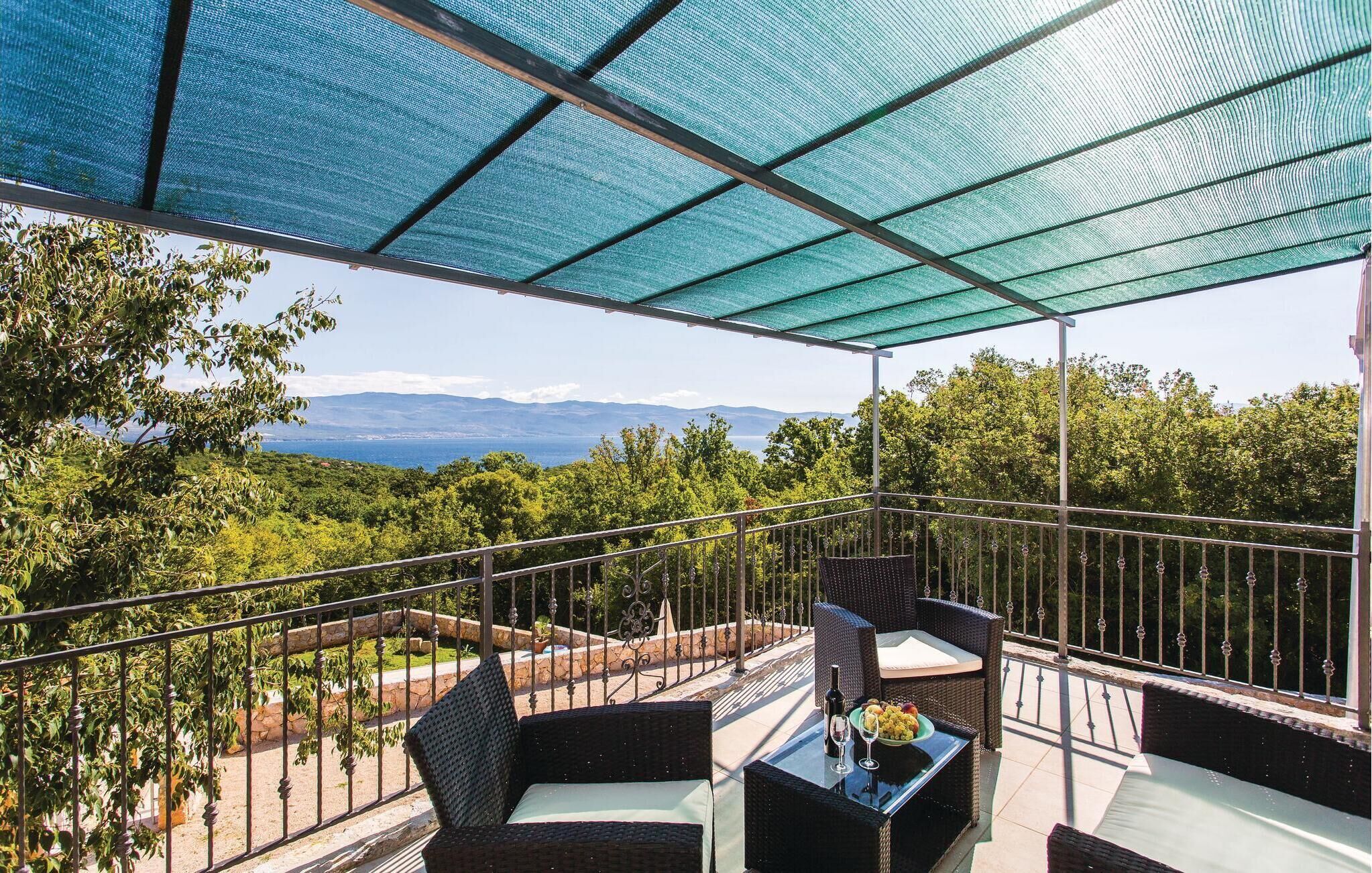A covered outdoor seating area with wicker furniture and a glass table, overlooking a scenic landscape with trees and mountains.