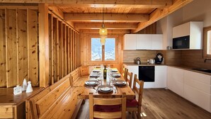 Interno di una baita in legno con un tavolo da pranzo per quattro persone, una cucina con mobili bianchi e una finestra con vista innevata.