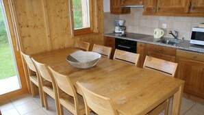 Une table à manger en bois avec des chaises, un grand saladier en osier et un coin cuisine avec des placards en bois, un four à micro-ondes et une machine à café.