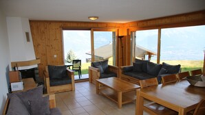 Un vaste espace de vie agrémenté de meubles en bois, de grandes fenêtres offrant une vue sur les montagnes et d’une cheminée.