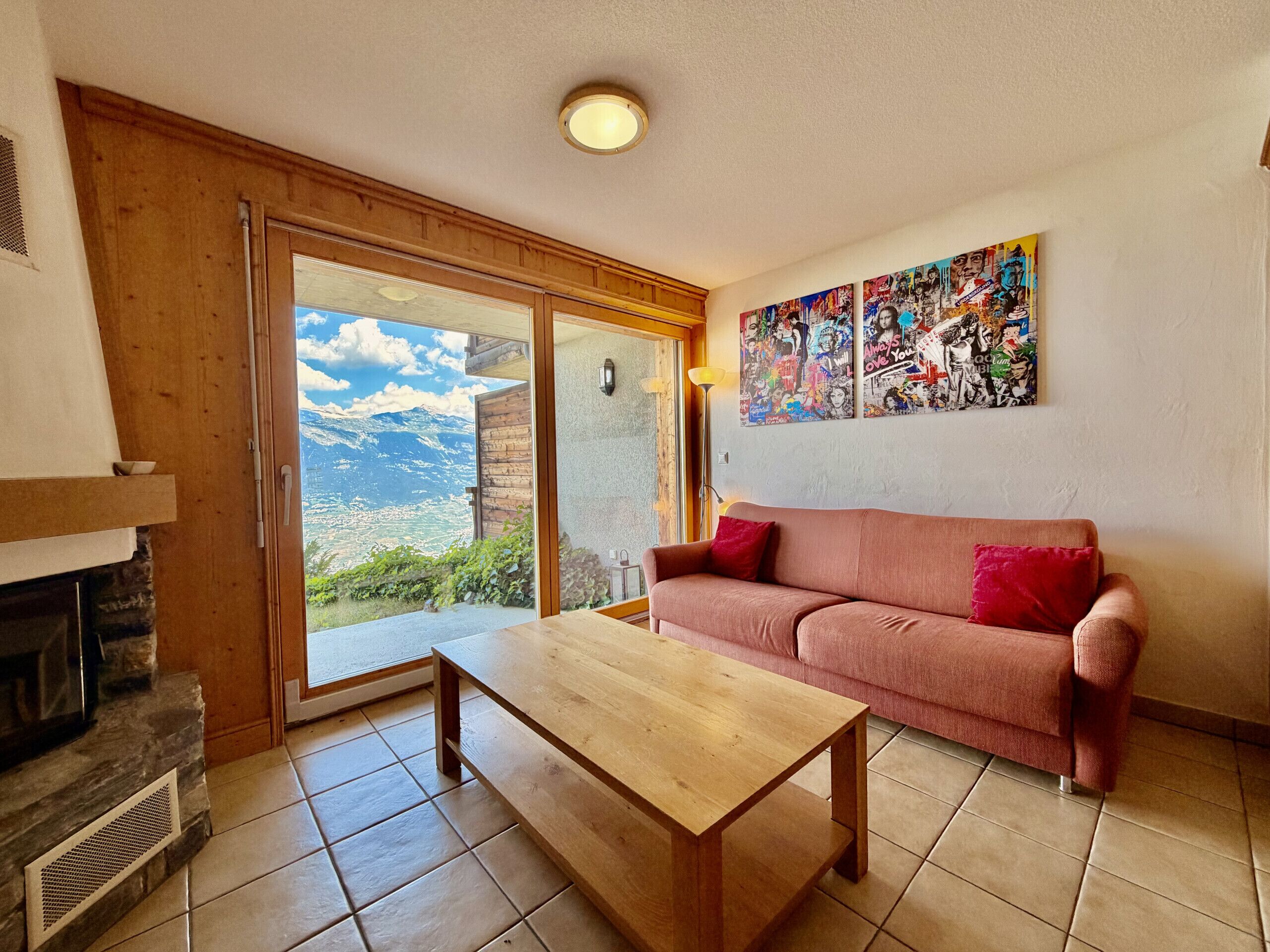 Un salon comprenant un canapé, une table basse, une cheminée, une grande fenêtre donnant sur les montagnes et un mur décoré de posters.