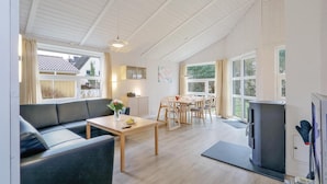 A spacious living area with a sofa, dining table, and chairs. There is a television and a fireplace. Large windows provide natural light and views of the outdoors. - Awesome home in Travemünde-Priwall (Travemünde-Priwall)