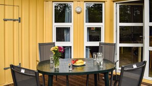 Ett runt glasbord med fyra stolar, en vas med blommor och en tallrik med frukt och glas på ett trädäck.