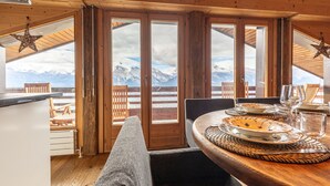 A wooden interior with a dining table set for two, a bench, and a view of snow-capped mountains through large glass doors.
