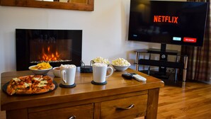 A living room with a fireplace, a TV displaying 'NETFLIX', and a wooden table with pizza, popcorn, and a coffee mug.