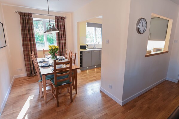 A dining area with a wooden table and chairs, a window with curtains, and a kitchen in the background.
