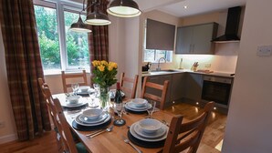 A dining area with a wooden table set for four, a vase with yellow flowers, and a kitchen in the background.