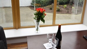 A room with a large window, a wooden table with a bottle and glasses, and a vase with flowers on the windowsill.