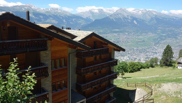 A multi-story wooden building with balconies, set against a backdrop of snow-capped mountains and a valley below.
