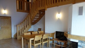 A wooden staircase leading to a loft area, a dining table with chairs, a television, and a stone fireplace.