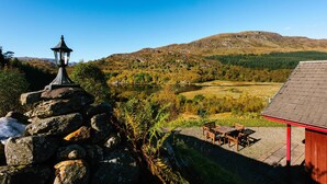 A stone lantern, a wooden table and chairs, and a red-roofed building with a view of a lake and hills. - Beautiful 5 star, 2 bedroom, 2 bathroom chalet with stunning views and wood burning stove (Crianlarich)