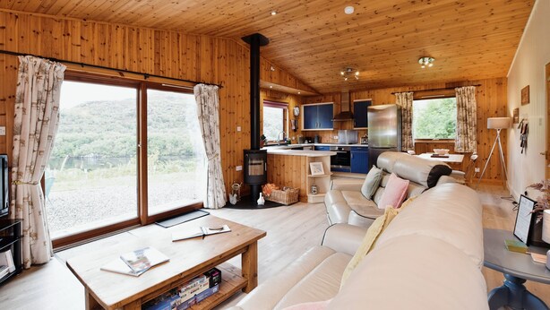 A cozy cabin interior with a wooden ceiling, a sofa, a dining table, a kitchen area, and large windows offering a view of the outdoors. - Beautiful 5 star, 2 bedroom, 2 bathroom chalet with stunning views and wood burning stove (Crianlarich)