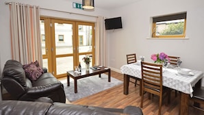 Ein Wohnzimmer mit einer Couch, einem Esstisch mit Stühlen, einem Couchtisch und einem an der Wand befestigten Fernseher.