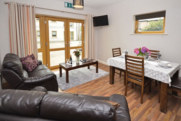 A living room with a sofa, a dining table with chairs, a coffee table, and a TV mounted on the wall. - Apartment 704 Letterfrack - sleeps 5 guests  in 3 bedrooms (Unknown)