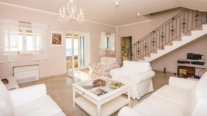 A spacious living room with white furniture, a chandelier, a staircase, and a view through an open door. - Cozy home in Brsecine (Brsecine)