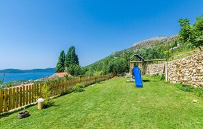 A grassy backyard with a wooden playhouse and a blue slide, a stone wall, and a wooden fence. - Cozy home in Brsecine (Brsecine)