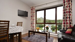 A living room with a dining table, a sofa, a TV mounted on the wall, and a large window with curtains. - Apartment 706 Letterfrack - sleeps 4 guests  in 3 bedrooms (Unknown)