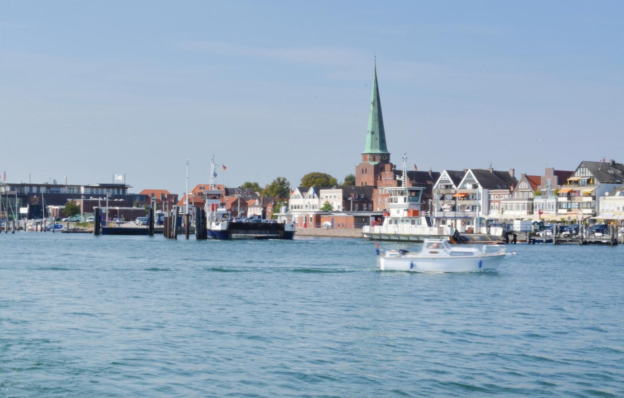 Un puerto con barcos, una iglesia con un campanario verde y edificios residenciales a lo largo del paseo marítimo.