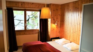 Une chambre avec un mur lambrissé en bois, un lit recouvert d’une couverture rouge, une lampe suspendue et une fenêtre avec des rideaux.