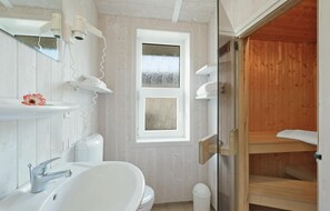 A bathroom with a white sink, a toilet, and a window with a view of trees. There is a wooden shelf with towels and a white trash bin. - Nice home in Travemünde-Priwall (Travemünde-Priwall)