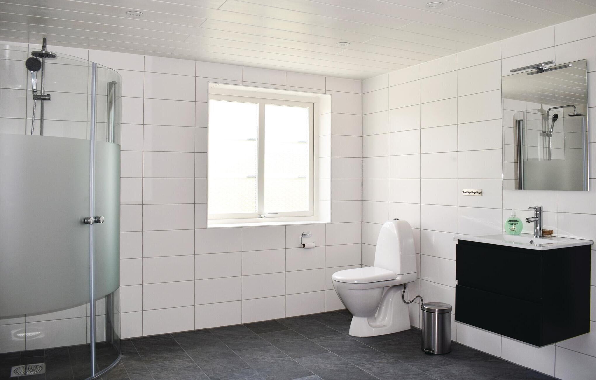 Une salle de bain moderne avec des carreaux blancs, une douche en verre, des toilettes, un lavabo avec un meuble noir et une fenêtre qui laisse entrer la lumière naturelle.