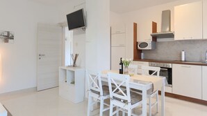 Eine moderne Küche mit weißen Schränken, einer eingebauten Mikrowelle und einem Herd. Es gibt einen Essbereich mit einem weißen Tisch und Stühlen sowie einen an der Wand befestigten Flachbildfernseher.